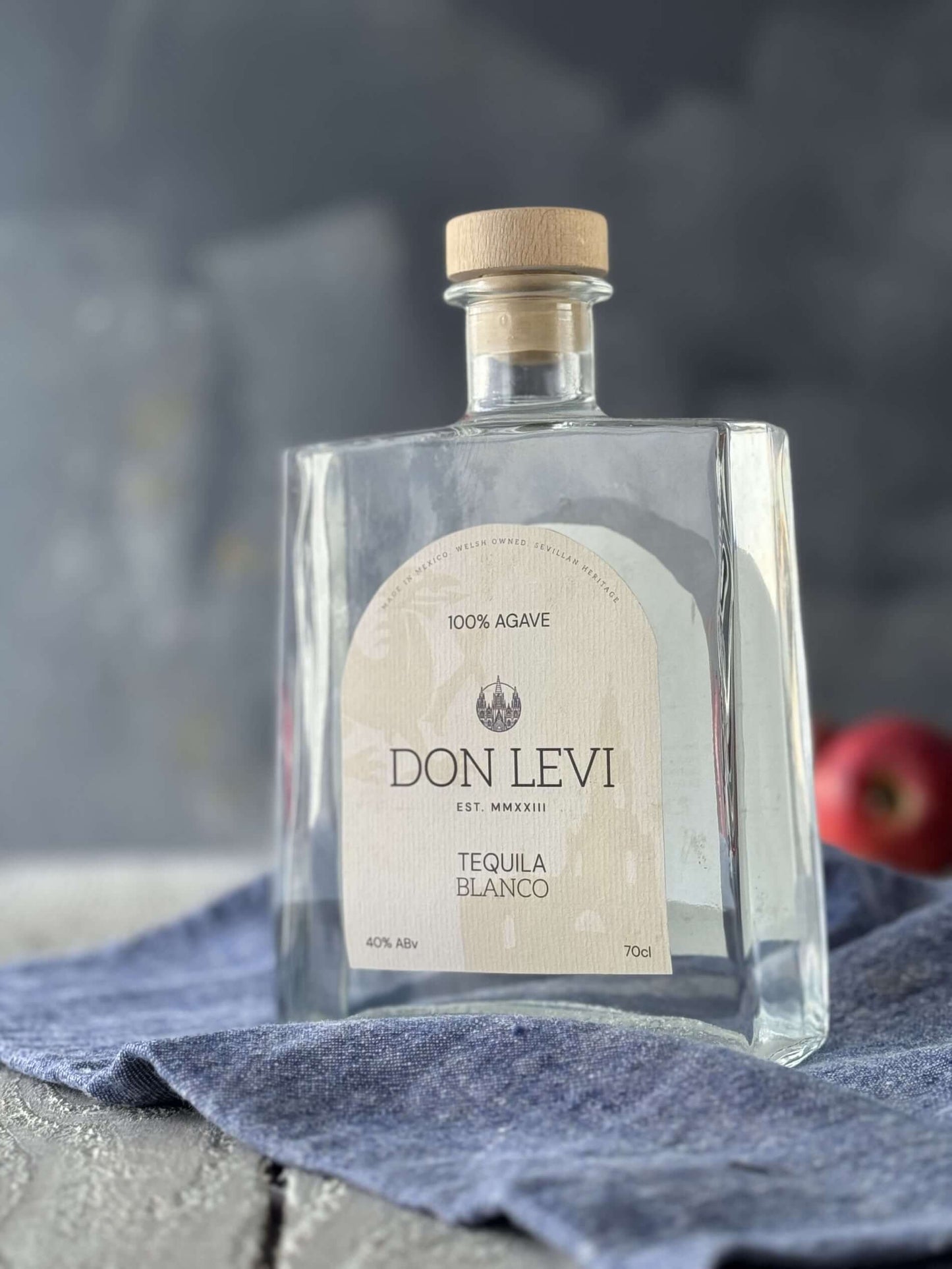 A clear glass bottle of Don Levi Tequila Blanco with a beige label and a light wood cap, resting on a blue cloth with a blurred dark background and a red apple in the distance.