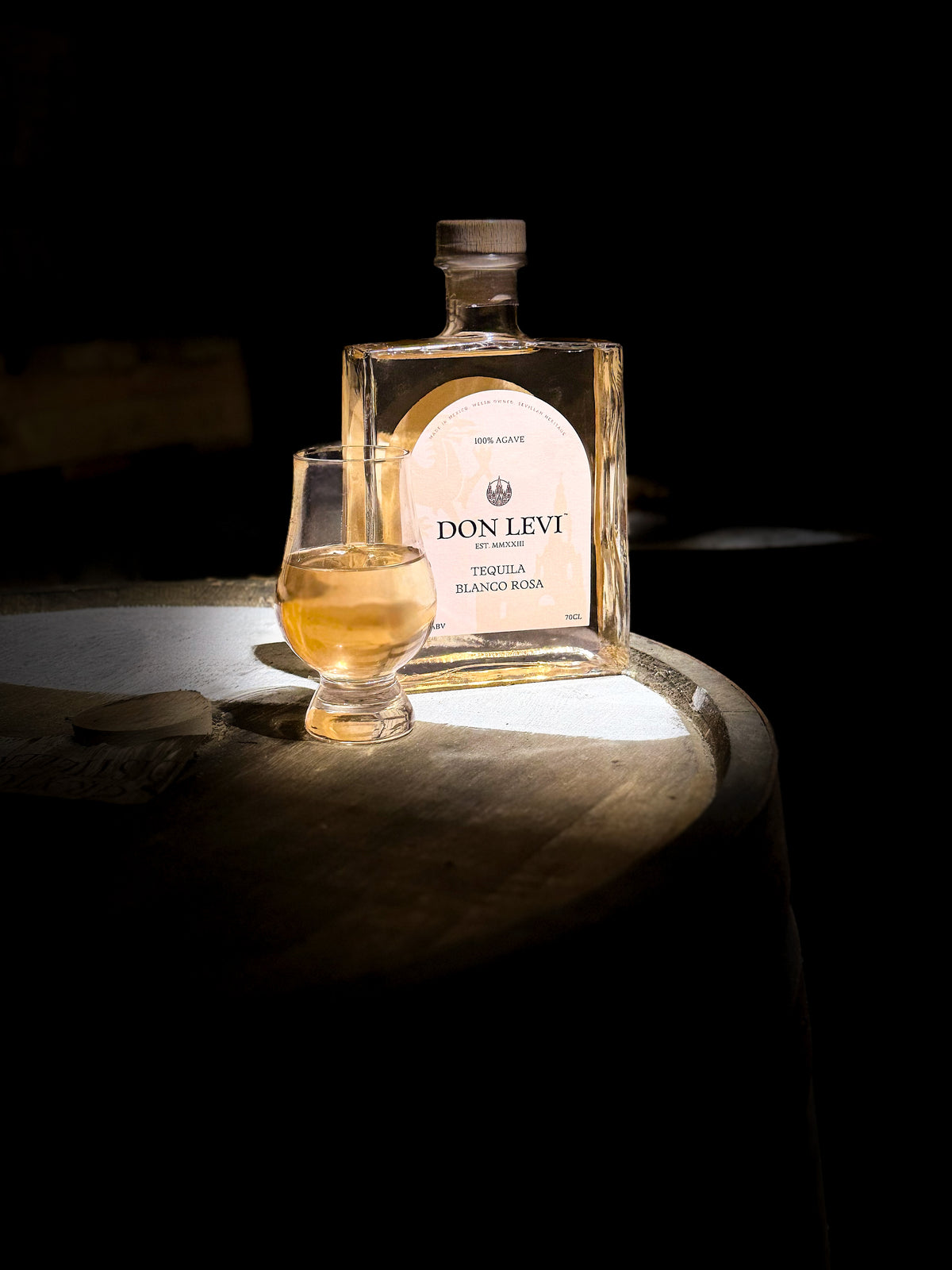 Bottle of Don Levi Rosa and a Glencairn glass with some Don Levi Rosa inside on a wooden barrel in a dim light room with a hit of light on the bottle and glass.