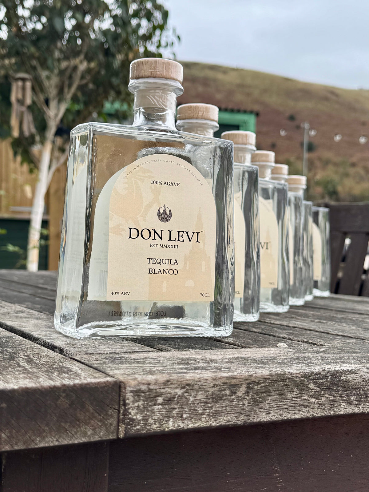 A row of Don Levi Tequila Blanco with cream labels and light wood caps, arranged on a weathered wooden table outdoors with hills and trees in the background.