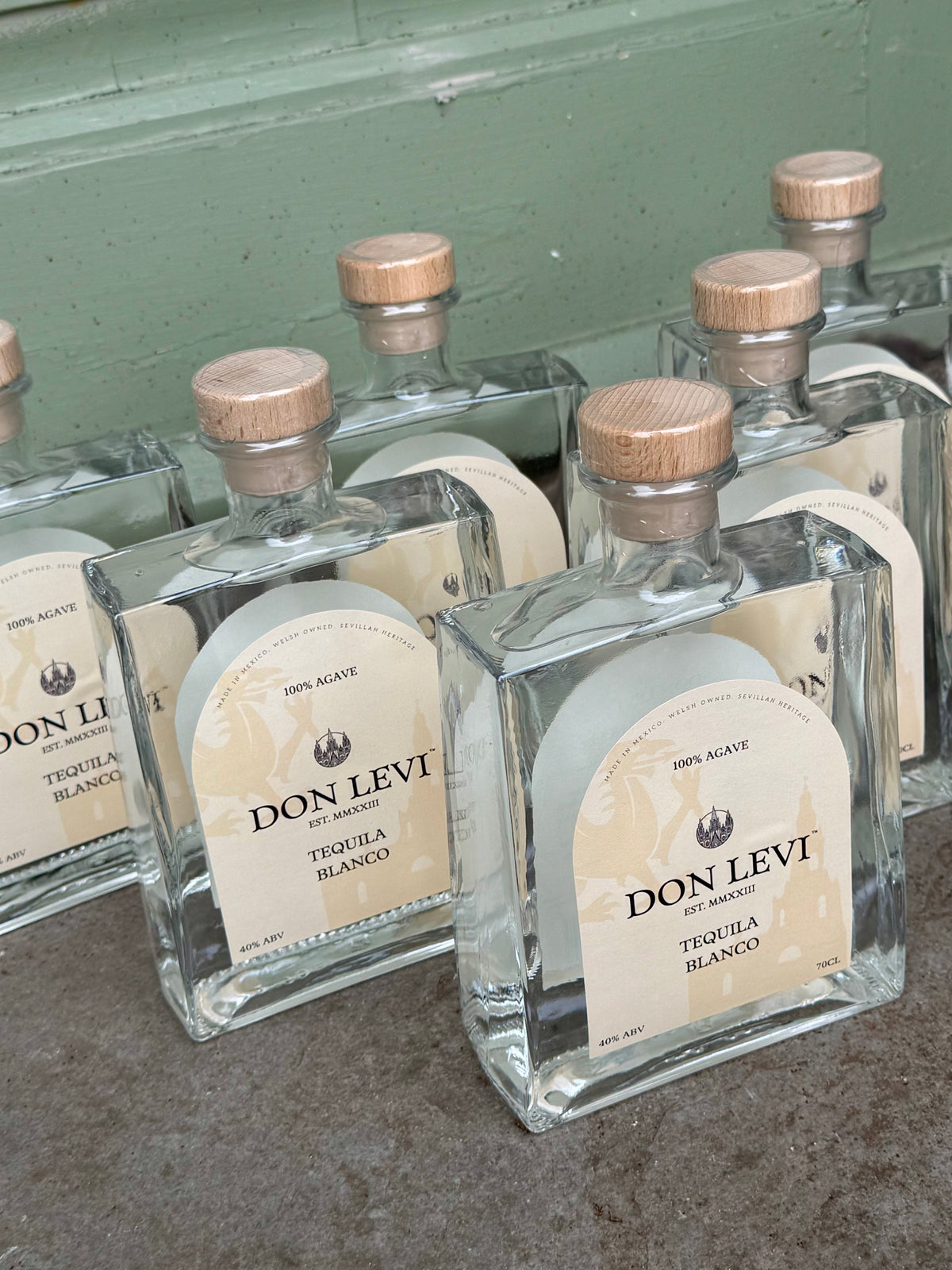 Several bottles of Don Levi Tequila Blanco with cream labels and light wood caps are arranged on a concrete surface against a painted green wall.