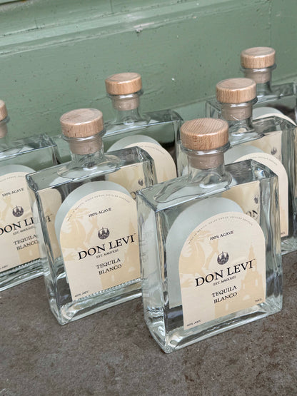 Several bottles of Don Levi Tequila Blanco with cream labels and light wood caps are arranged on a concrete surface against a painted green wall.
