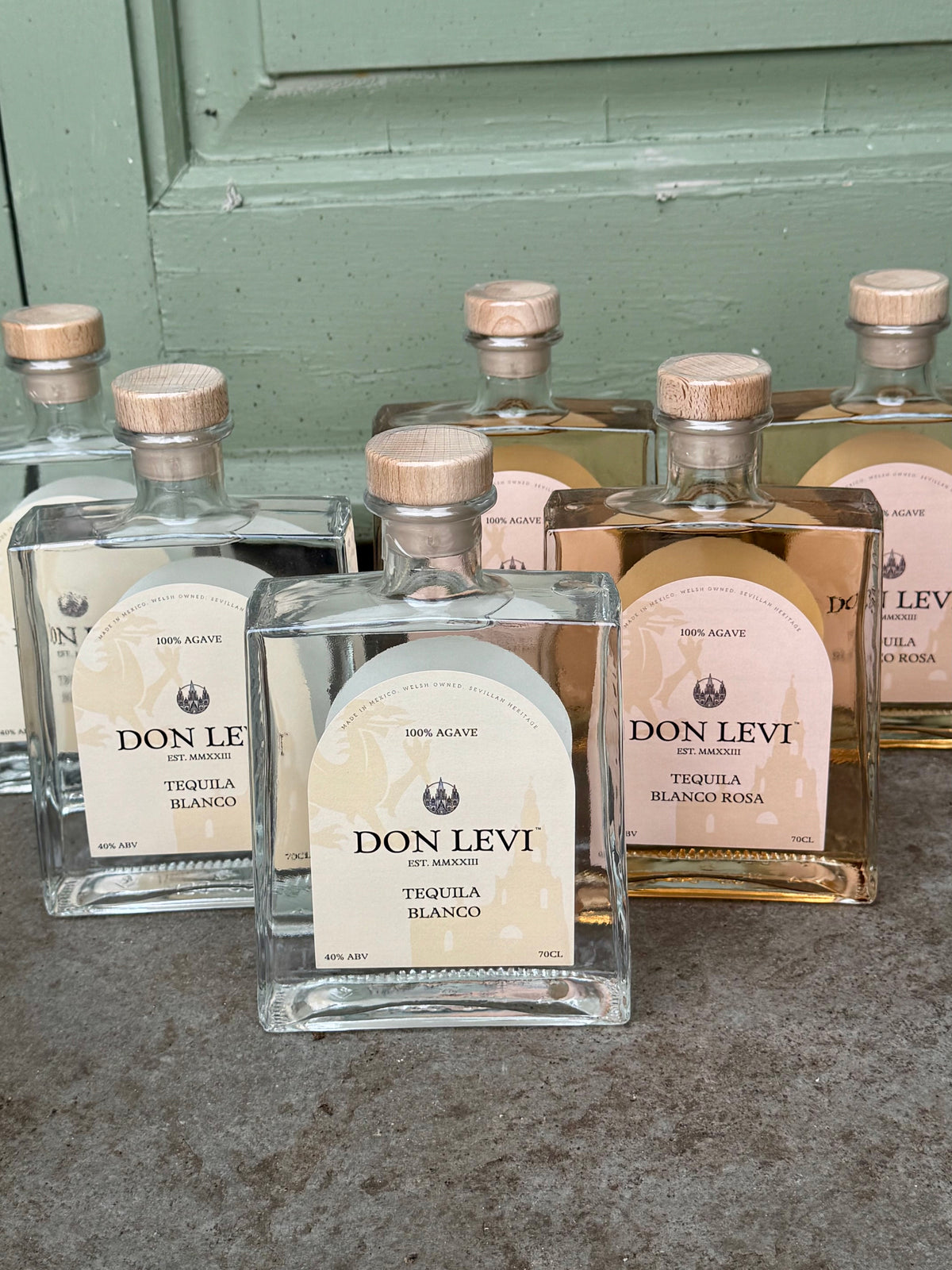 Several bottles of Don Levi tequila (blanco & rosa) arranged on a gray surface in front of a green wooden door. 