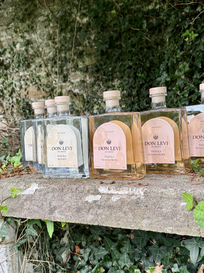 Several of Don Levi blanco & rosa  tequila are lined up on a stone ledge outdoors, with green ivy and foliage in the background.