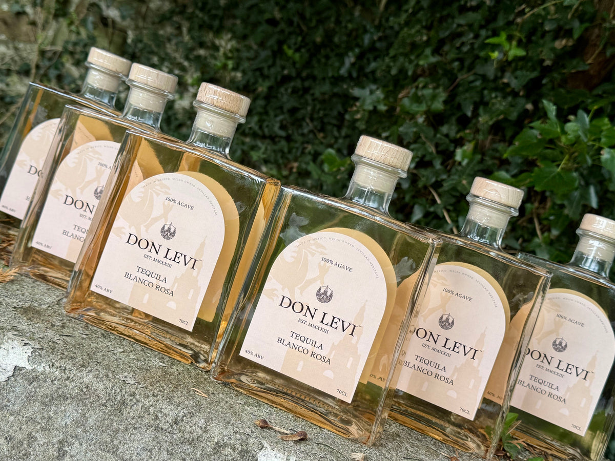 Several clear glass bottles of Don Levi Tequila Rosa with cream labels and light wood caps, arranged on a stone surface with green foliage in the background.