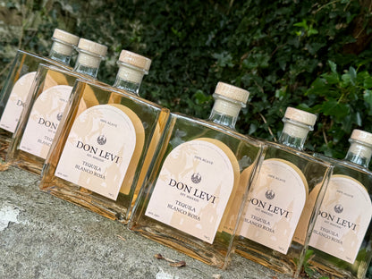 Several clear glass bottles of Don Levi Tequila Rosa with cream labels and light wood caps, arranged on a stone surface with green foliage in the background.