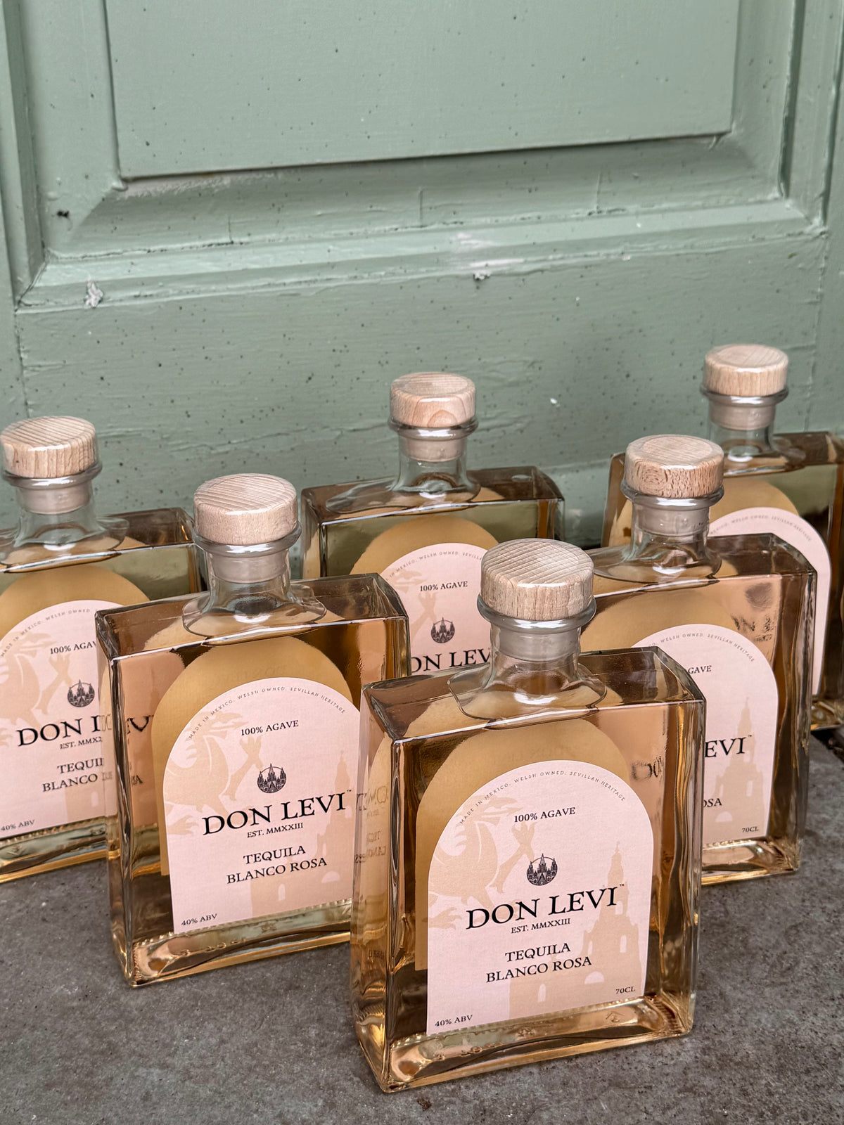 Several clear glass bottles of Don Levi Tequila Blanco Rosa with pale pink labels and cork stoppers, arranged on a gray surface in front of a muted green paneled door.