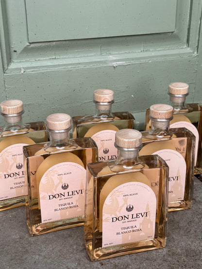 Several clear glass bottles of Don Levi Tequila Blanco Rosa with pale pink labels and cork stoppers, arranged on a gray surface in front of a muted green paneled door.