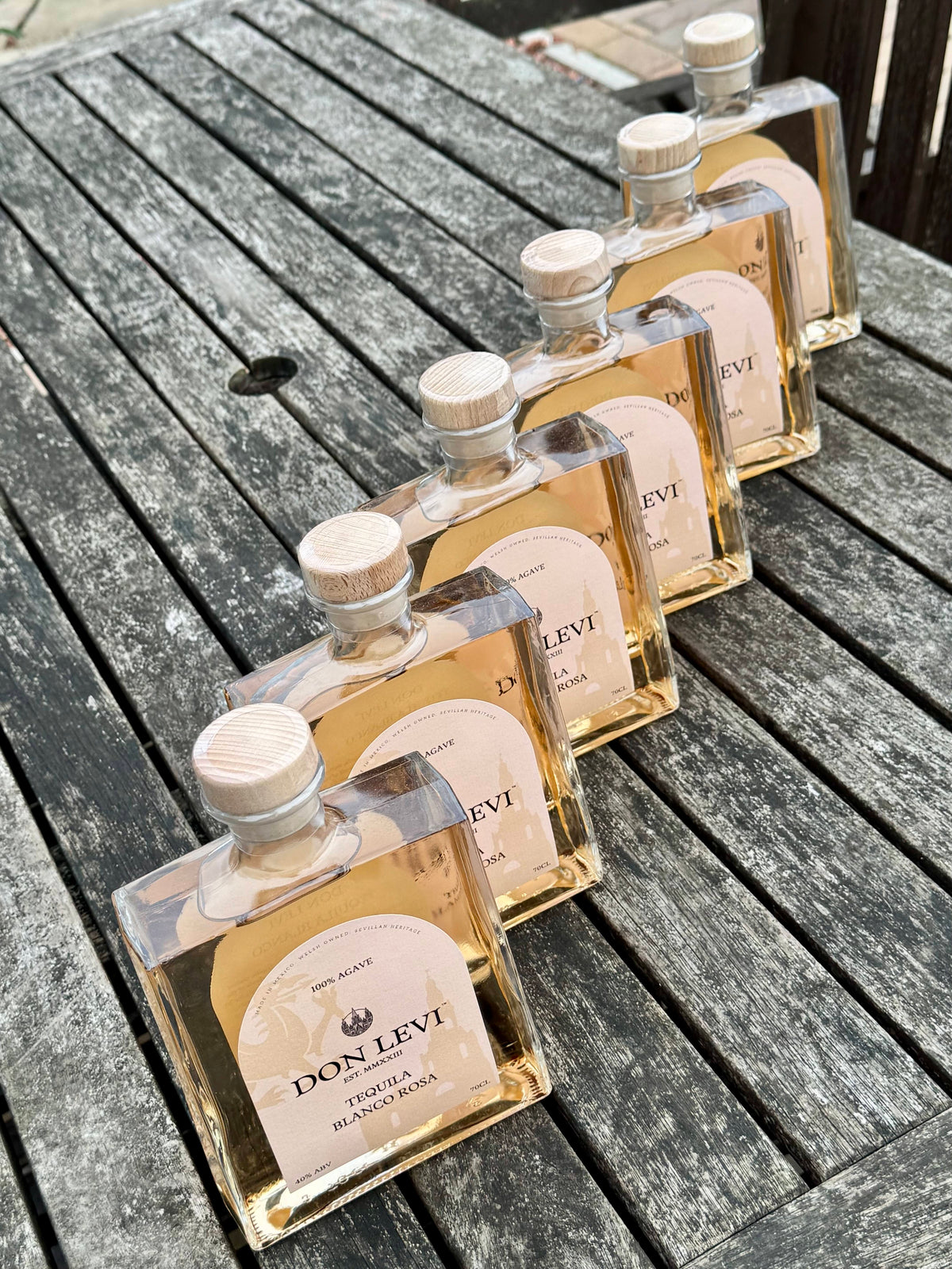 6 clear glass bottles of Don Levi Rosa tequila arranged diagonally on a weathered gray wooden table.
