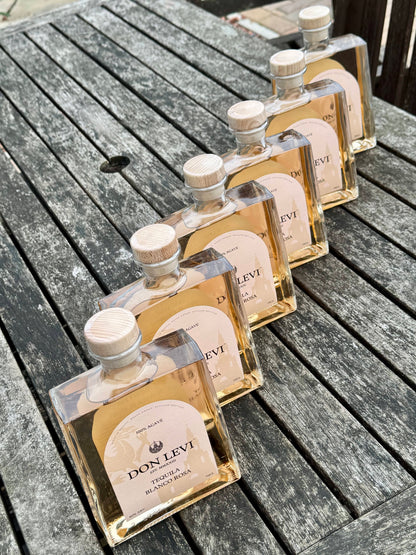 6 clear glass bottles of Don Levi Rosa tequila arranged diagonally on a weathered gray wooden table.