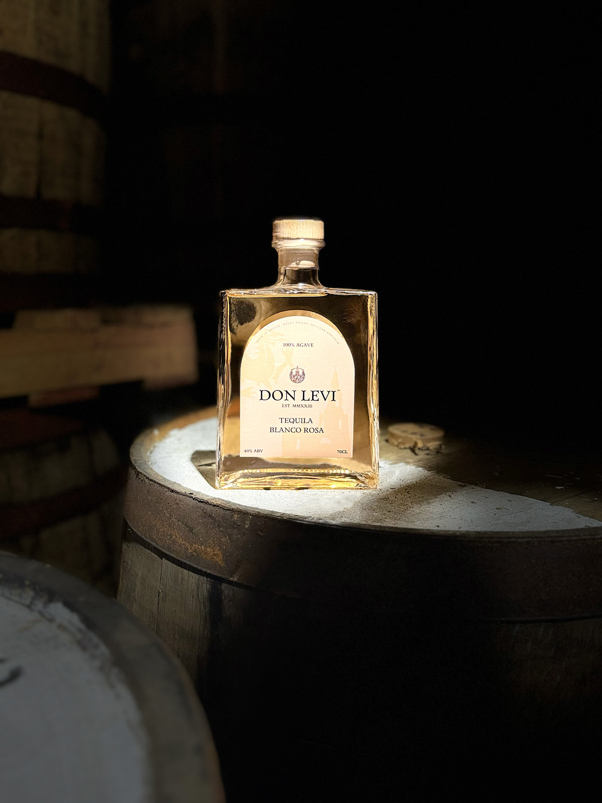 A bottle of Don Levi Rosa Tequila on a barrel in a dim lit room, with some light coming from the left brighting the bottle and barrel.