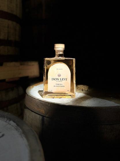 A bottle of Don Levi Rosa Tequila on a barrel in a dim lit room, with some light coming from the left brighting the bottle and barrel.