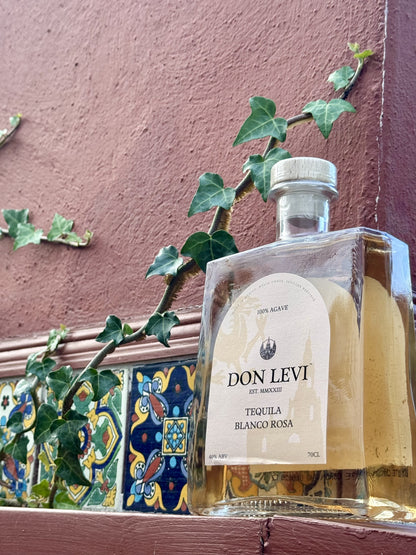 A bottle of Don Levi Rosa Tequila set against a red terracotta wall with green ivy and colourful mosaic tiles.