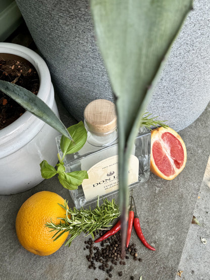 A bottle of Don Levi Blanco next to some agave plants, citrus fruits, peppercorns, peppers and herbs. Highlighting the tasting noes.